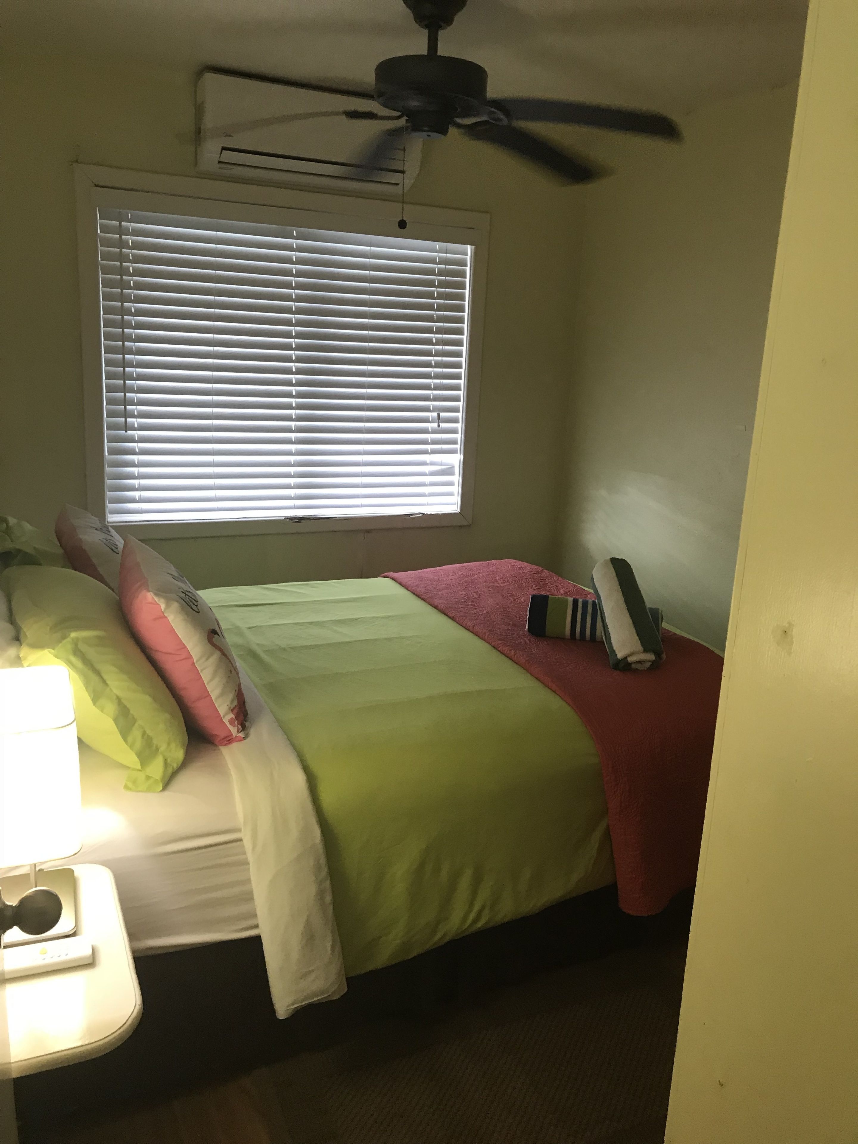 Ein Schlafzimmer mit einem Bett, Kissen, einem Nachttisch mit Lampe, einem Deckenventilator und einem Fenster mit Jalousien.