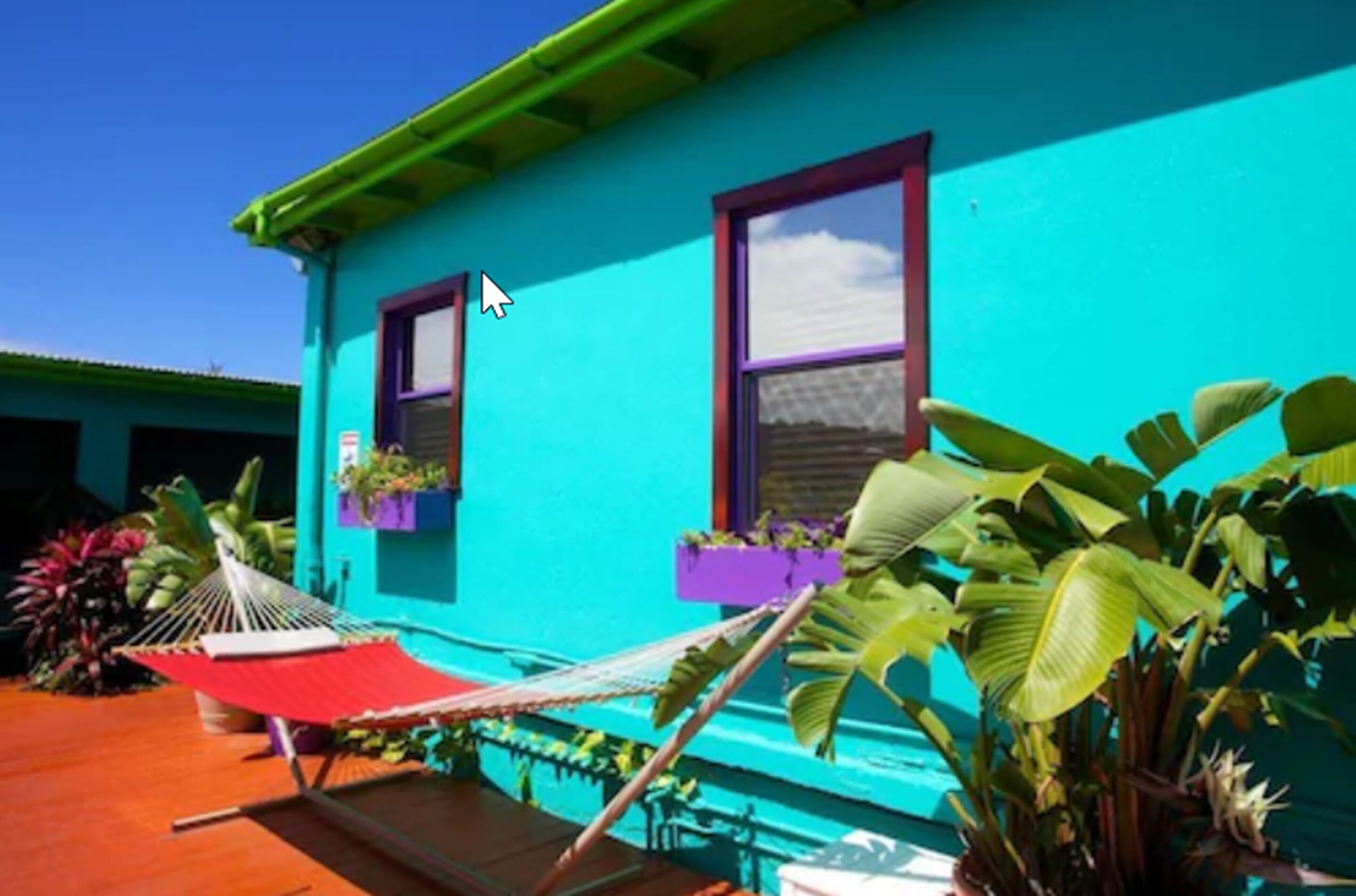 A turquoise building with two windows, a red hammock, and potted plants.