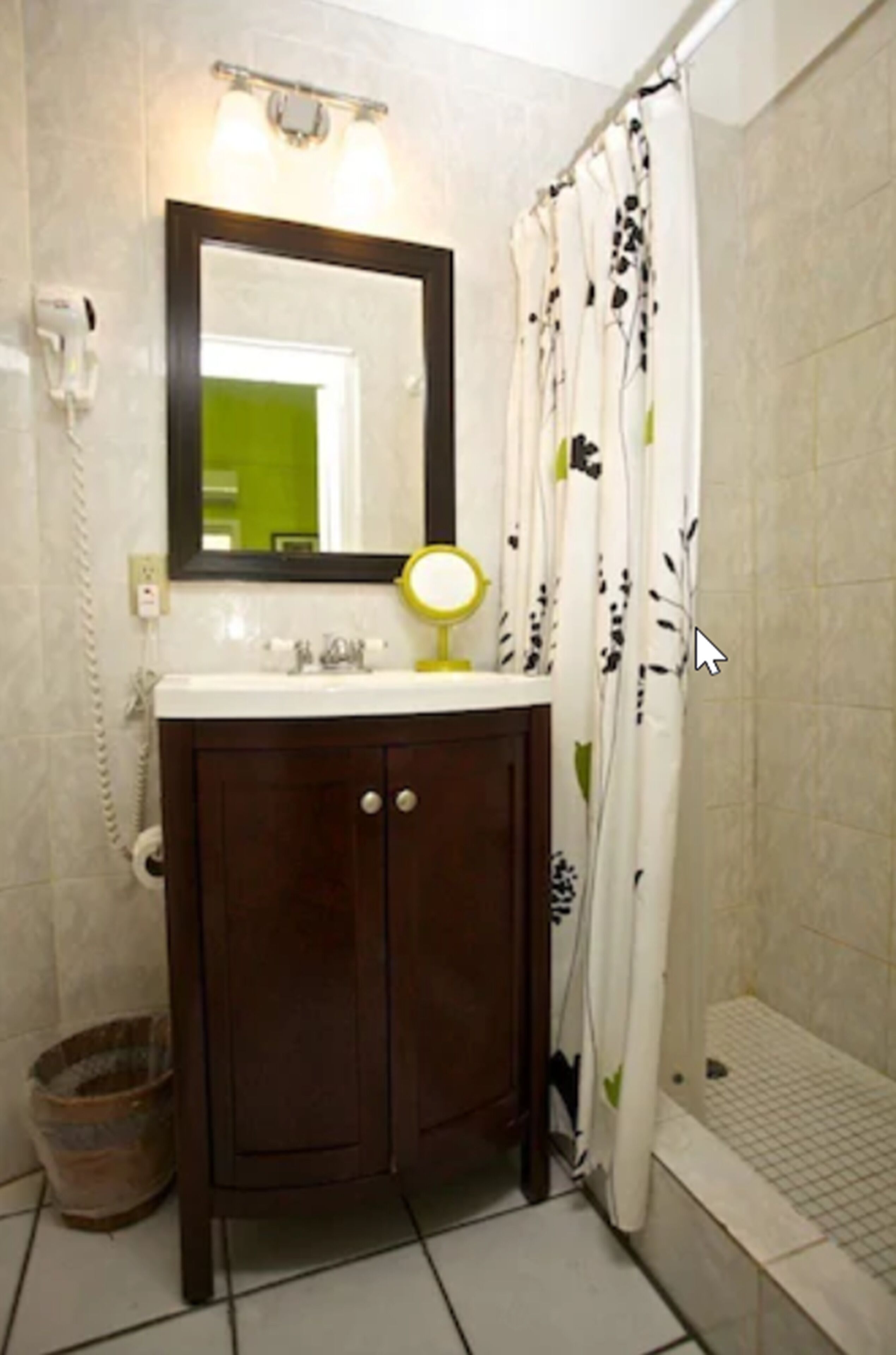 A bathroom with a white sink, dark wooden cabinet, a mirror with a yellow light, a white shower curtain with black and green patterns, and a white toilet.