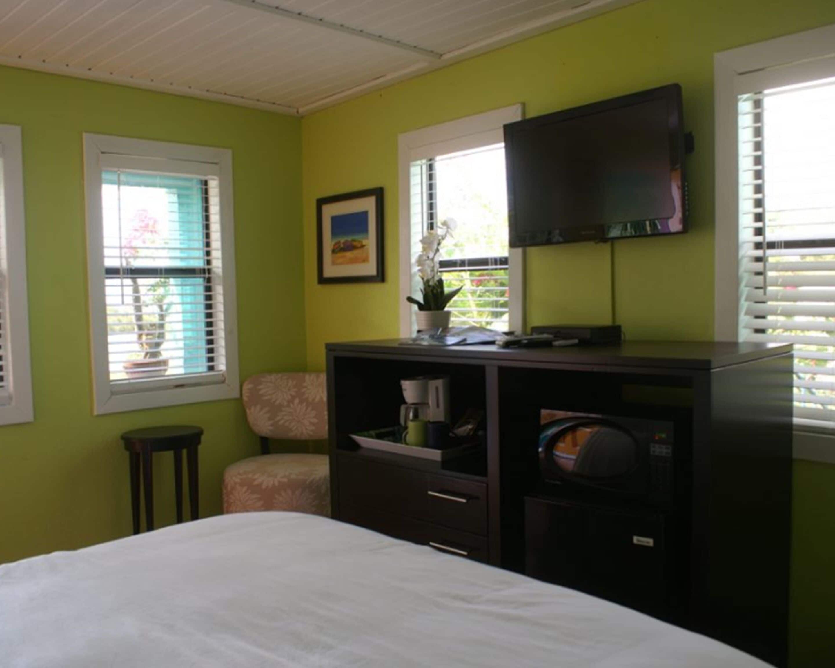 A bedroom with a bed, a desk with a flat-screen TV, a chair, and a window with blinds.