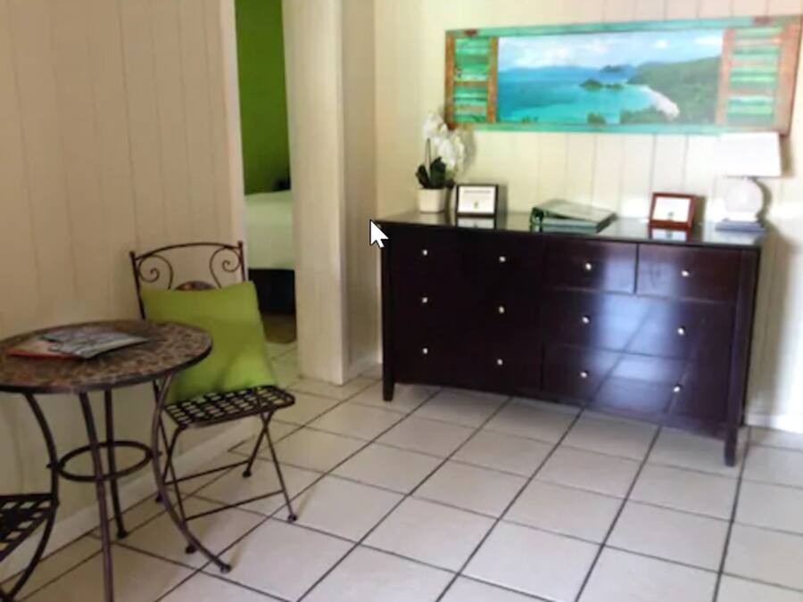 A room with a small round table, a green chair, a dark-colored dresser, and a framed picture on the dresser.