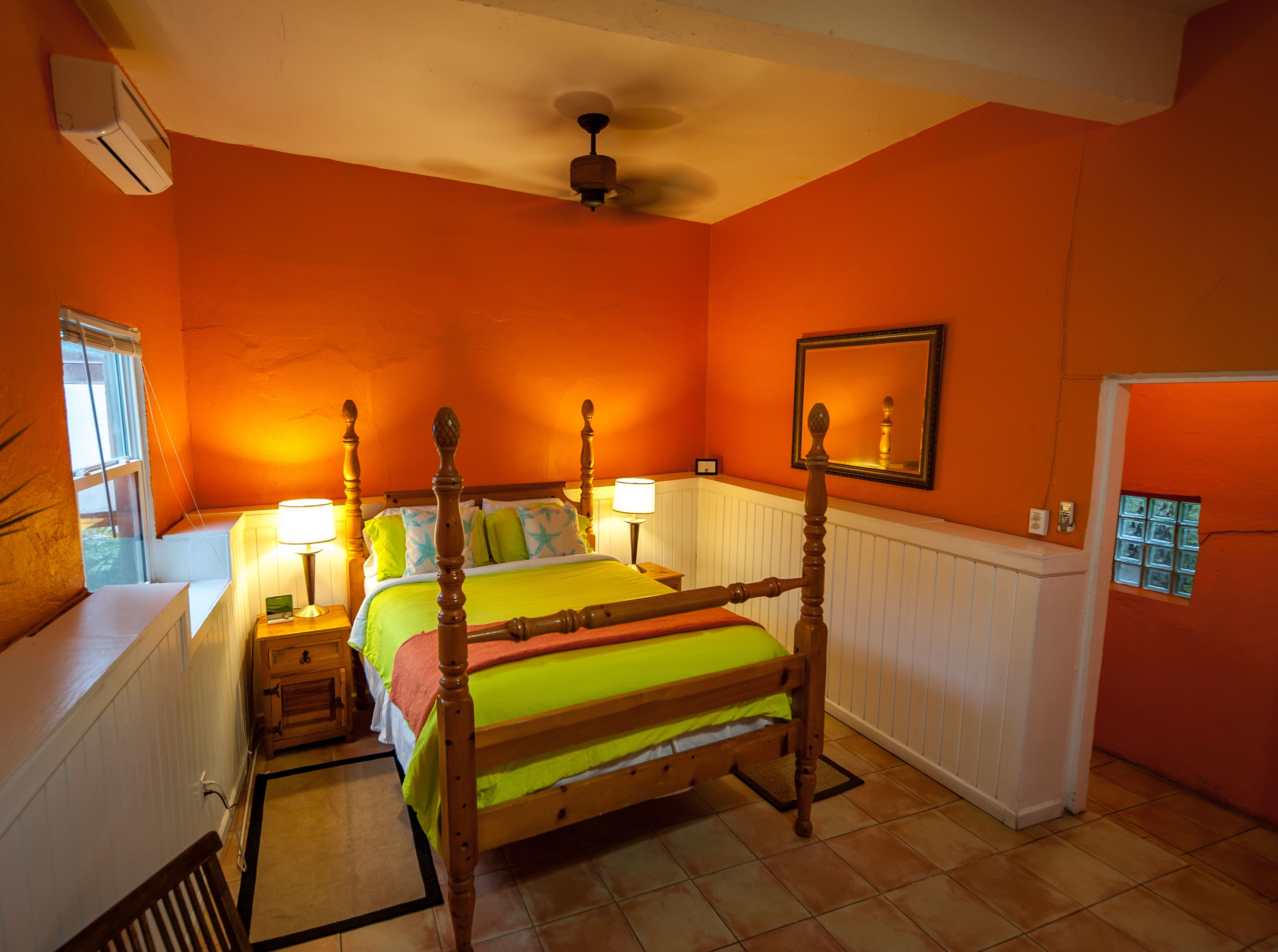 A bedroom with a four-poster bed, two bedside lamps, a ceiling fan, a window with blinds, and a door with a small window.