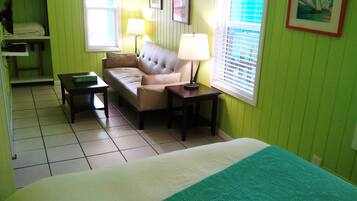Una habitación con cama, sofá, mesa y lámpara. Tiene ventanas con persianas, un cuadro en la pared y una pared verde.
