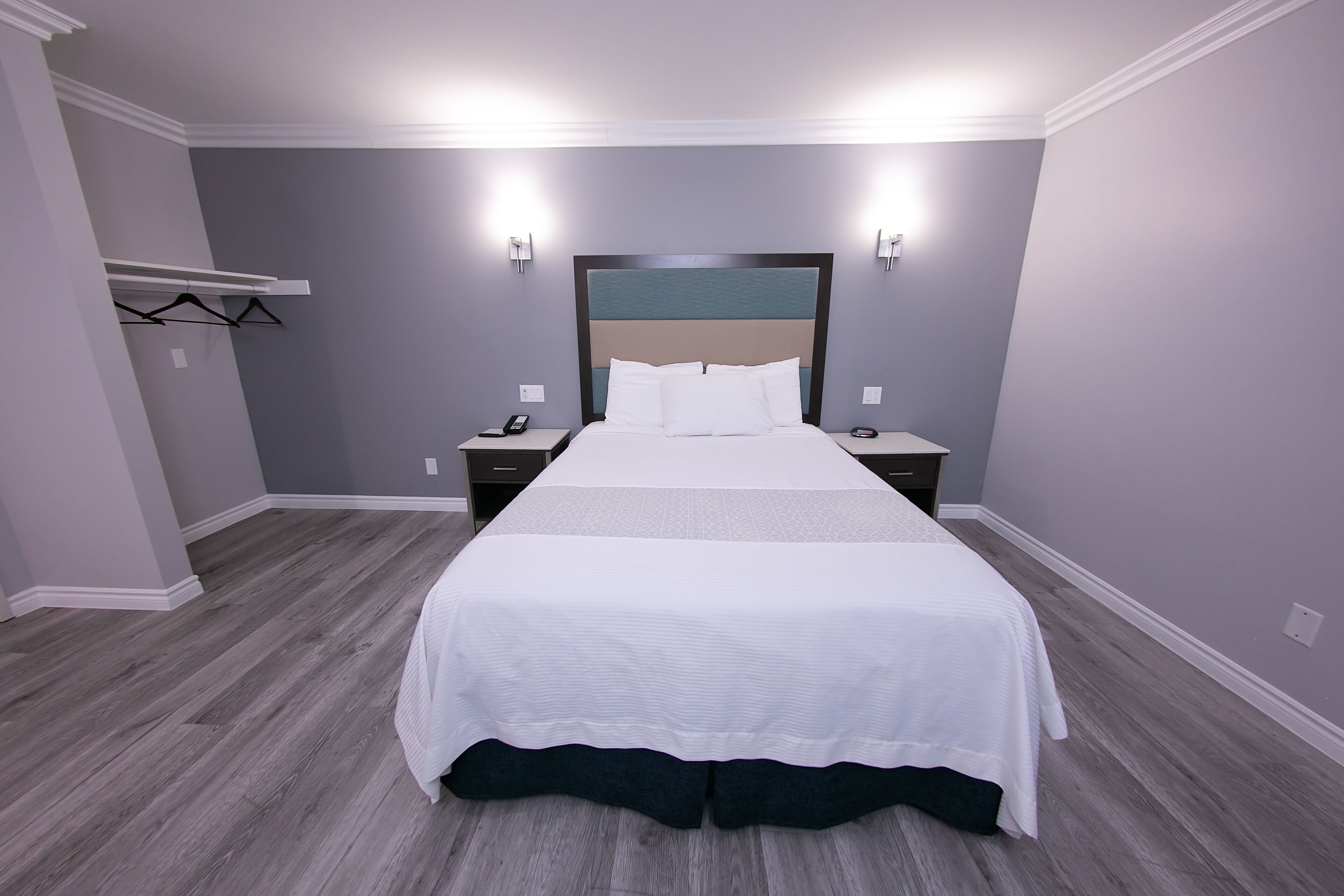 A neatly made bed in a hotel room with a grey wall, a wooden floor, and a headboard with a blue panel.