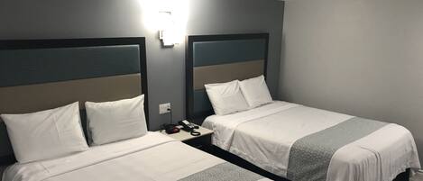 Deux lits jumeaux avec du linge de lit blanc et une couvre-lit à motifs géométriques, une table de chevet avec un téléphone et une prise électrique, et un luminaire fixé au mur.