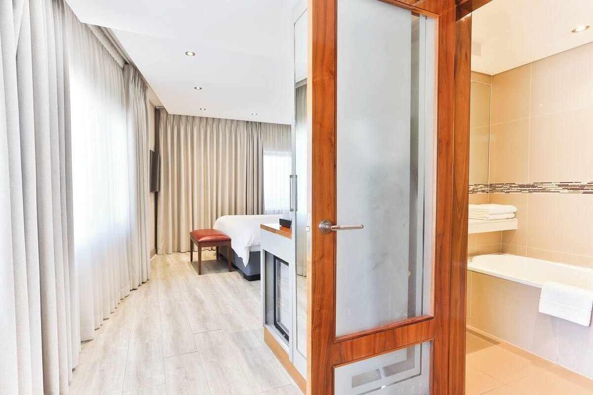 Ein modernes Hotelzimmer mit Holztür, einem Bett, einem Schreibtisch, einem Stuhl, einem Fernseher und einem Badezimmer mit Badewanne und Handtuchhalter.