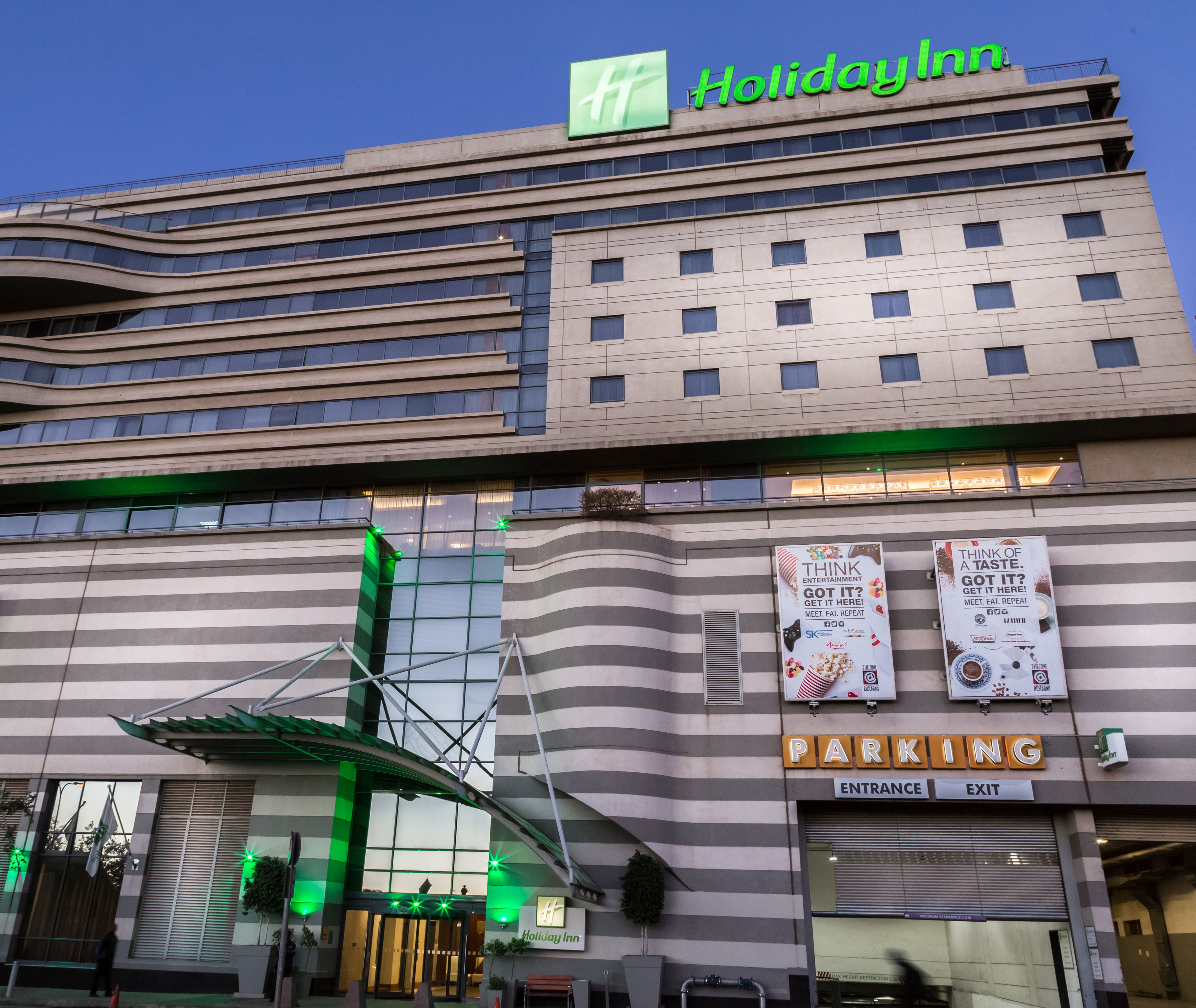 A Holiday Inn hotel with a modern design, featuring a prominent green logo, a parking sign, and multiple banners advertising entertainment options.