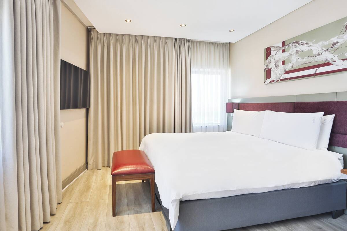 Ein Hotelzimmer mit einem großen Bett, einer roten Bank, einem Flachbildfernseher und einem modernen abstrakten Gemälde an der Wand.