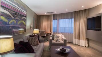 A hotel room with a sofa, armchair, TV, and a painting on the wall.