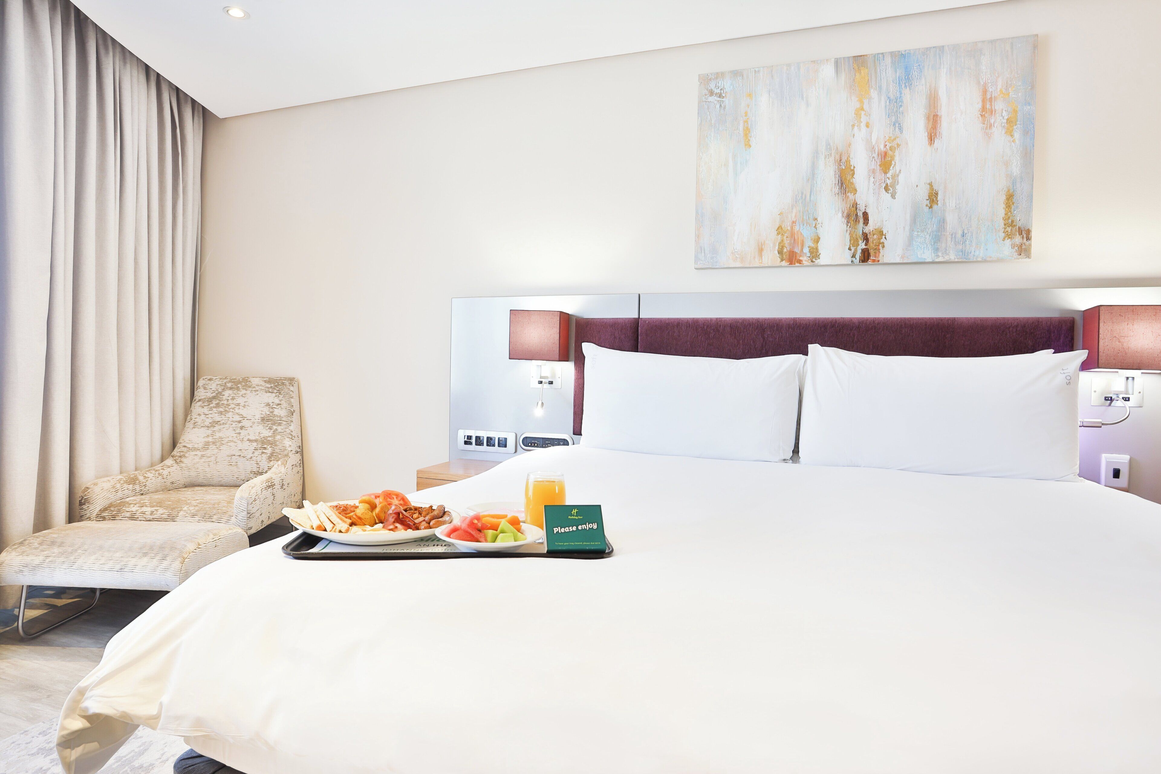 A hotel room with a bed, a chair, a tray with food, and a painting on the wall.