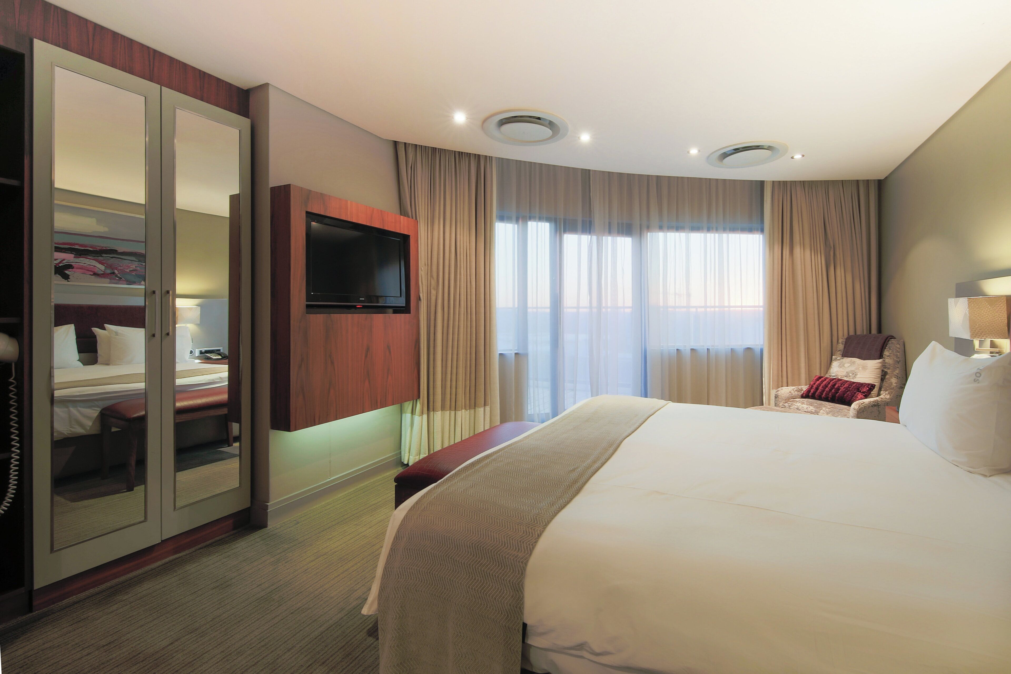 A hotel room with a large bed, a TV mounted on the wall, a wardrobe, a bench, and a chair.