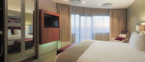 A hotel room with a large bed, a TV mounted on the wall, a wardrobe, a bench, and a chair.