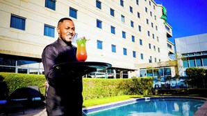 A hotel with a pool, a person holding a tray with a drink, and a building with multiple windows. - Holiday Inn Johannesburg - Rosebank by IHG (Johannesburg)