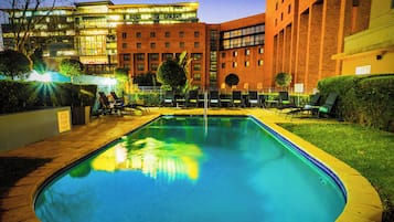 Une piscine entourée de chaises longues, elle-même bordée d’une pelouse bien entretenue et d’un bâtiment à plusieurs étages doté de grandes fenêtres.