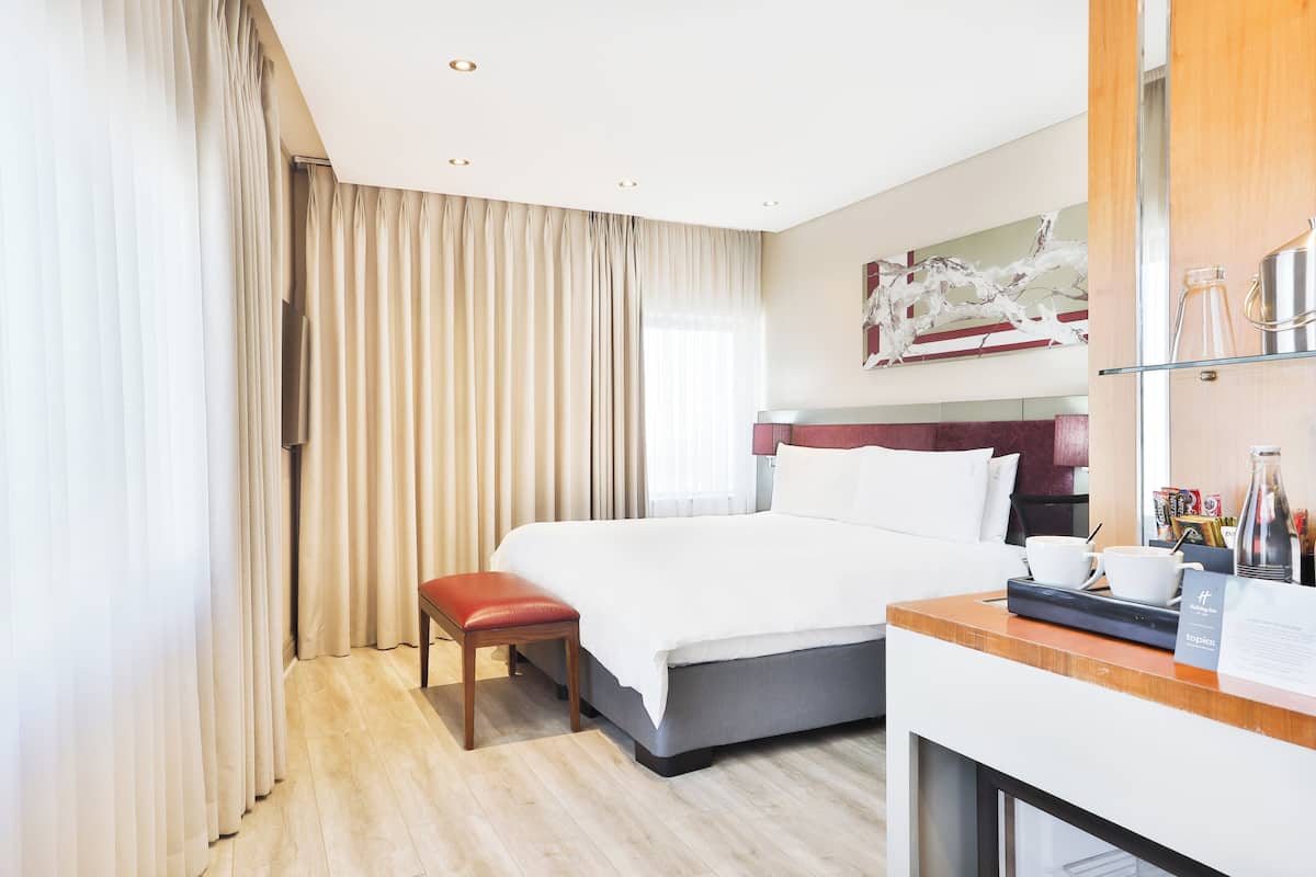 Ein Hotelzimmer mit Bett, Schreibtisch, Stuhl, einem Fenster mit Vorhängen und einem Bild an der Wand.