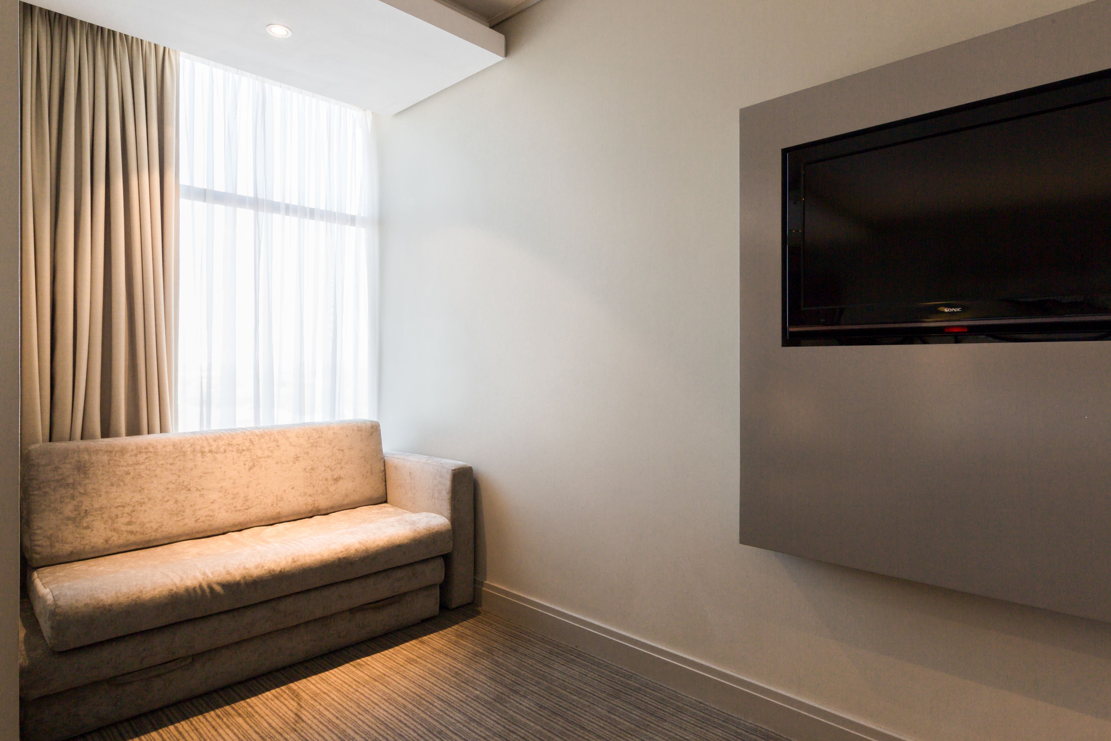 A modern hotel room with a beige sofa, a large flat-screen TV mounted on the wall, and a large window with sheer curtains.