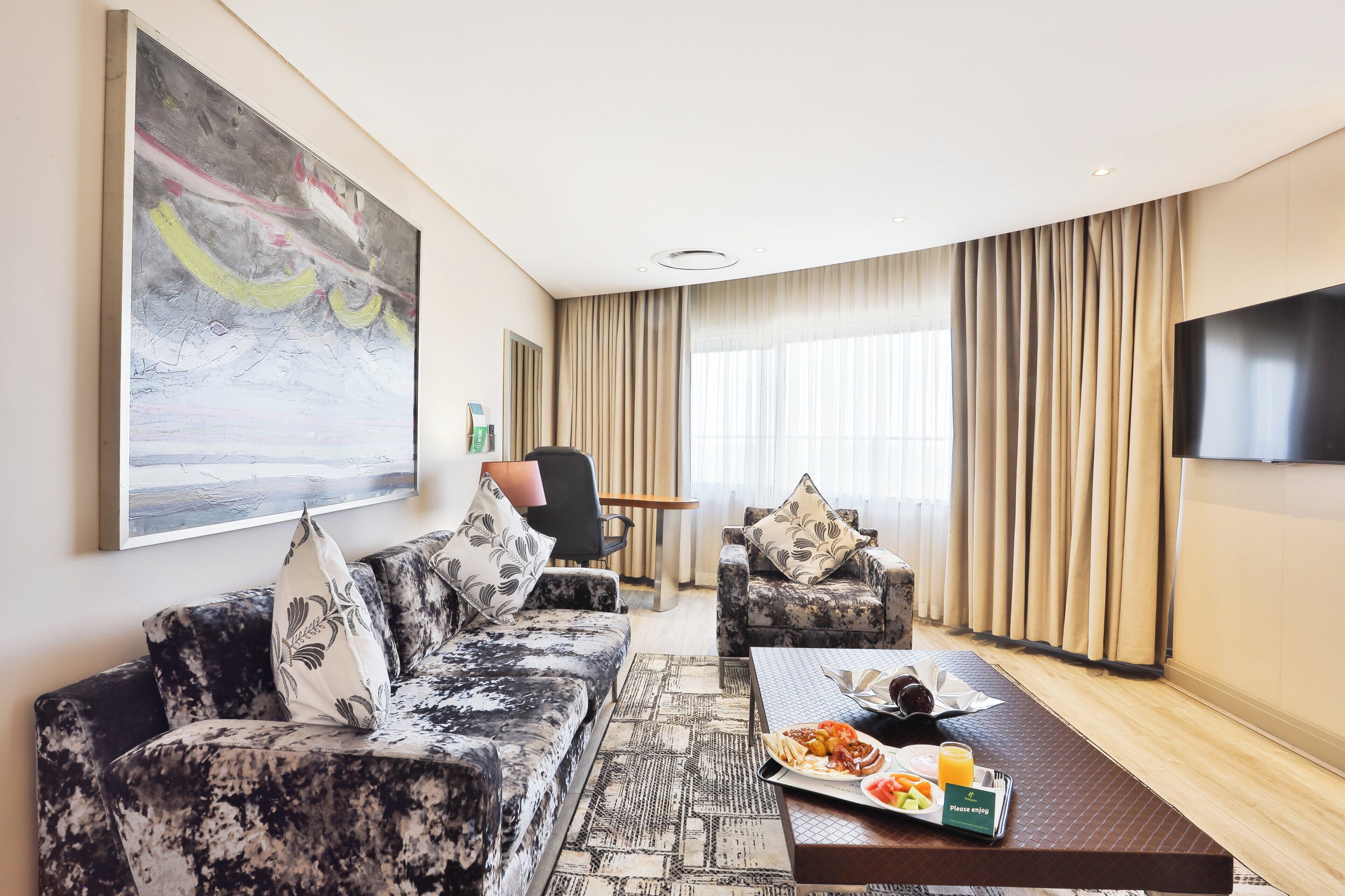 A modern hotel room with a sofa, armchair, dining table, and a large TV.
