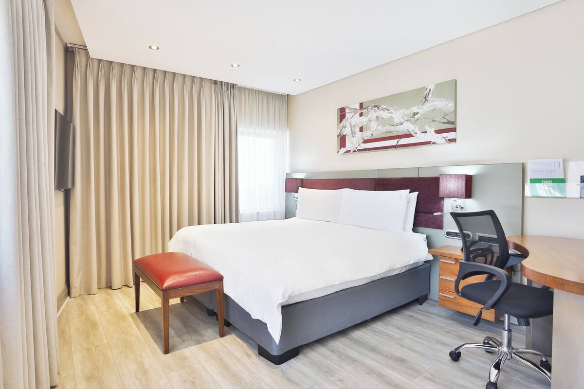 Ein Hotelzimmer mit einem großen Bett, einem Schreibtisch mit Stuhl, einem Fernseher und einem Bild an der Wand.