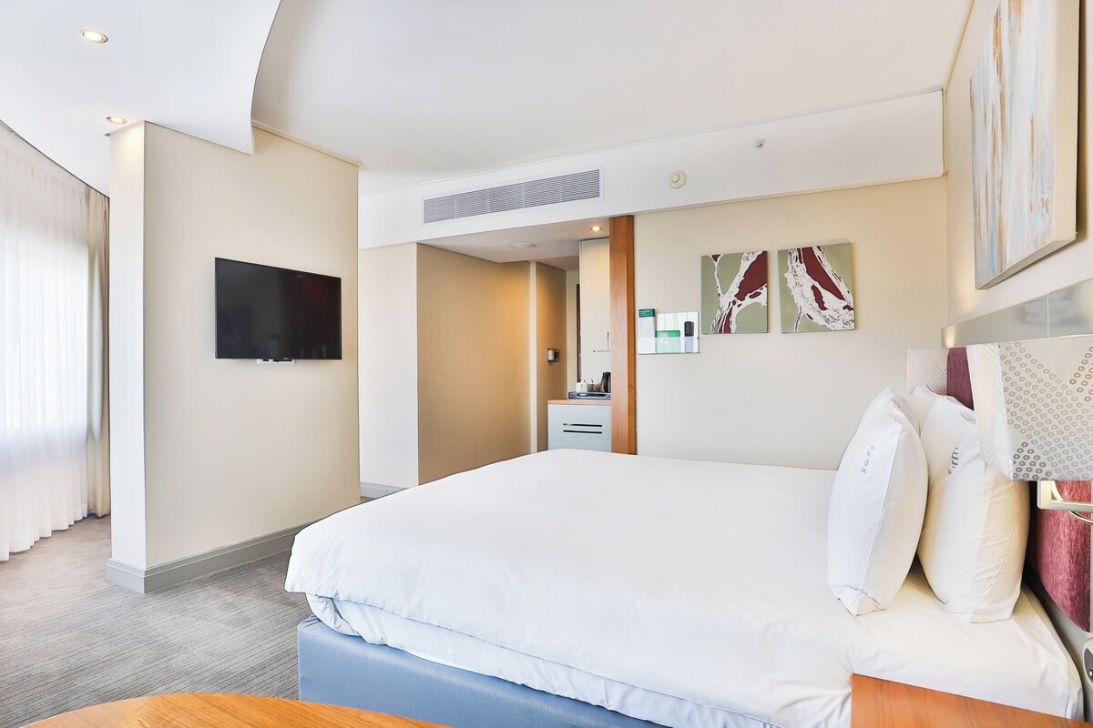 Ein Hotelzimmer mit einem großen Bett, einem flachbildfernseher an der Wand, einem Schreibtisch mit Stuhl und Kunstwerken an der Wand.