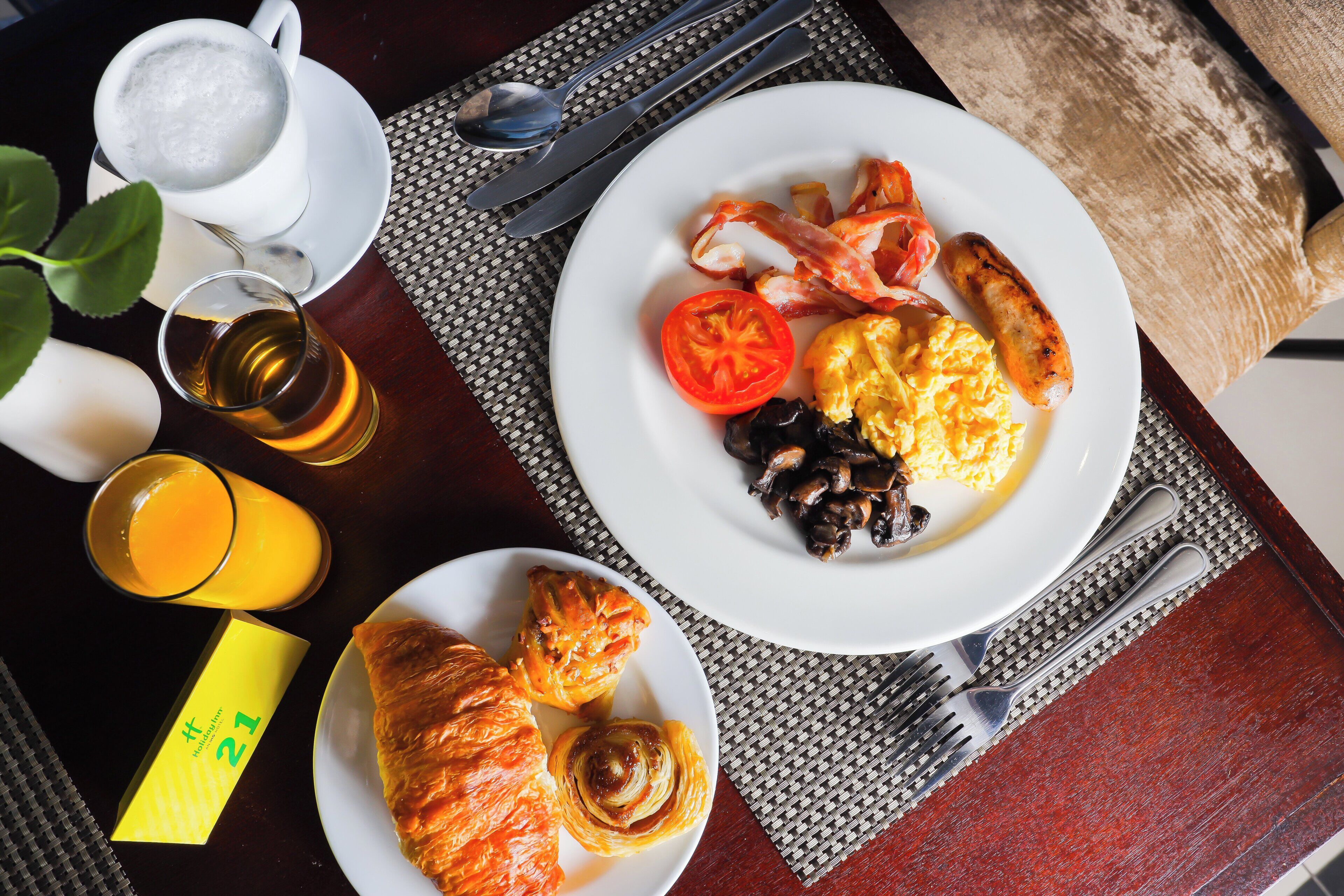 A breakfast setting with a plate of eggs, bacon, sausage, and a tomato on a table with a napkin, cutlery, and a glass of orange juice.