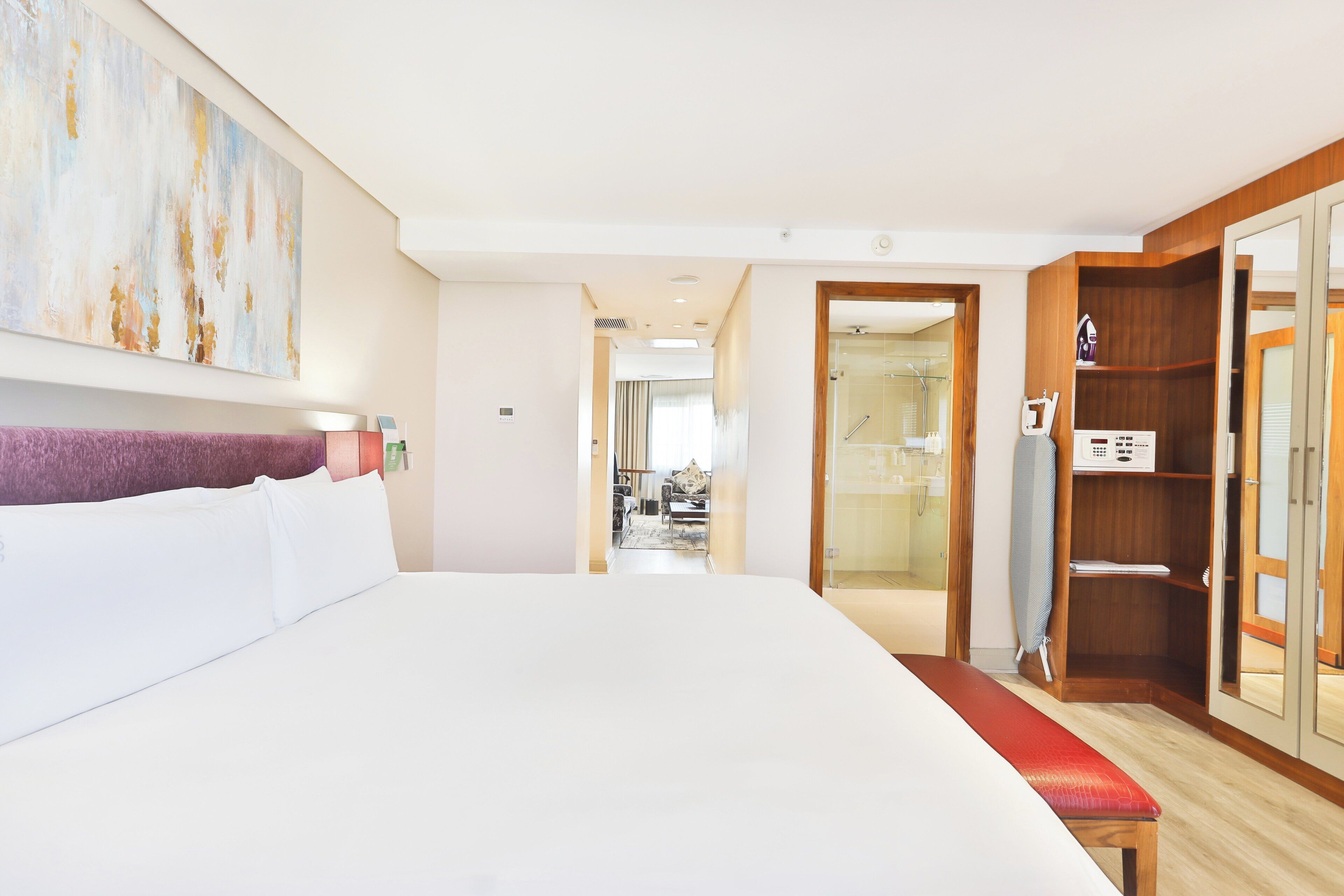 A hotel room with a large bed, a wooden wardrobe, a bench, and a view into the bathroom.