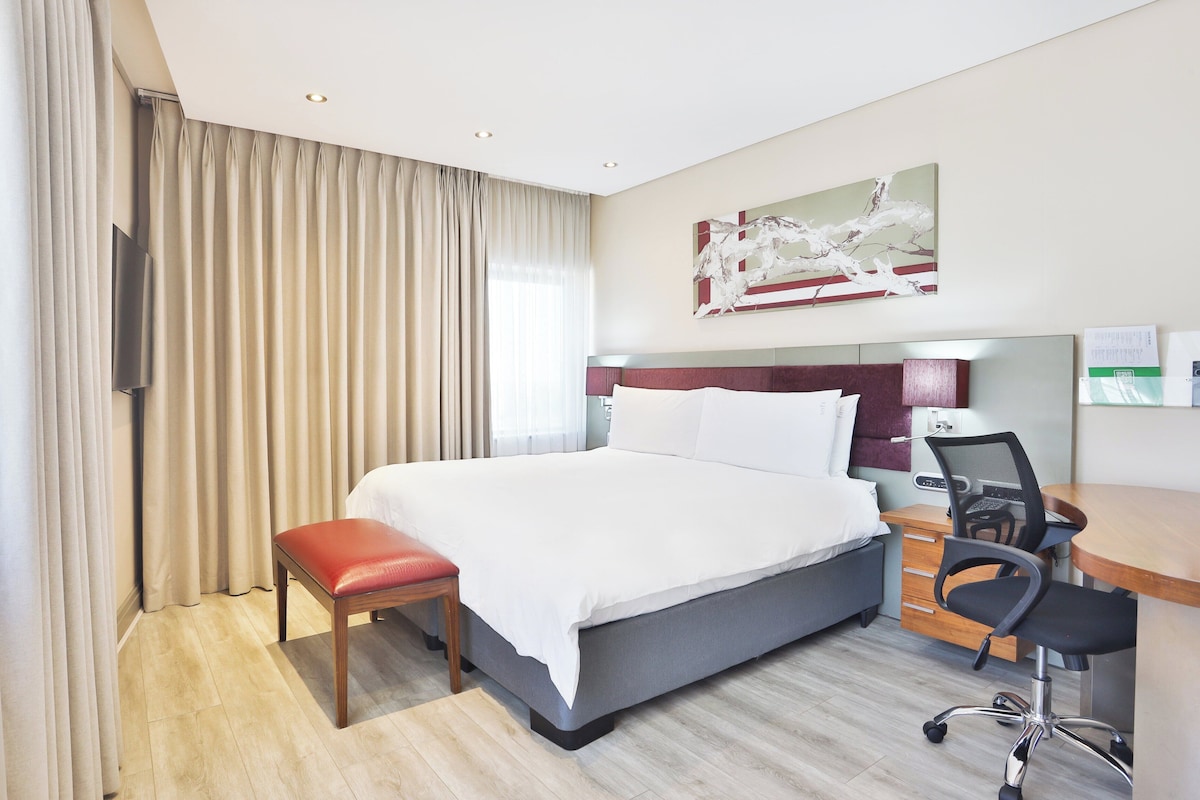 A hotel room with a large bed, a desk with a chair, a television, and a painting on the wall.