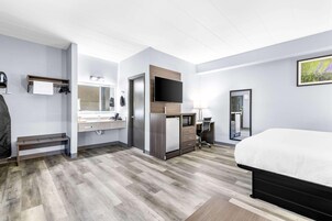 A hotel room with a bed, a desk, a chair, a TV, a mirror, and a bathroom with a sink and towel rack.