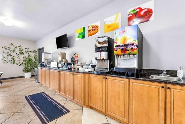 A modern kitchen with wooden cabinets, a coffee machine, a juice dispenser, and a flat-screen TV mounted on the wall.