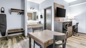 Een moderne hotelkamer met een houten eettafel, een aan de muur bevestigde televisie, een bureau met stoel en een spiegel boven het wastafeltje zonder spiegel.