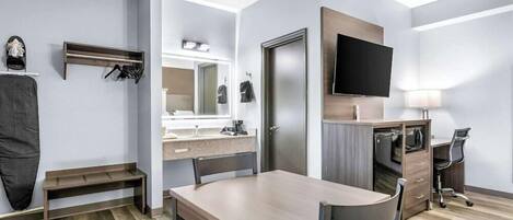 A modern hotel room with a wooden dining table, a TV mounted on the wall, a desk with a chair, and a mirror above the mirrorless vanity.