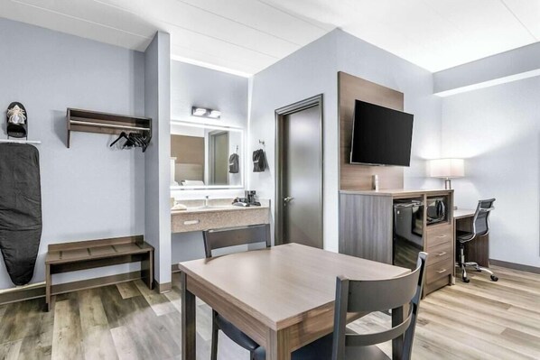 A modern hotel room with a wooden dining table, a TV mounted on the wall, a desk with a chair, and a mirror above the mirrorless vanity. - Americas Best Value Inn (Russellville)