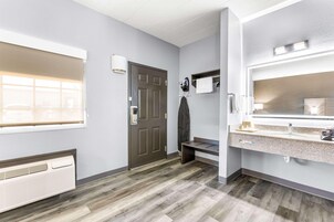 A hotel room with a wooden floor, a large window with blinds, a door, a bench, a vanity with a mirror, and a wall-mounted heater.