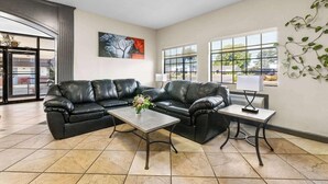 A spacious living room with a black leather sofa, a coffee table, and a lamp. There are large windows providing natural light and a view of the outside.