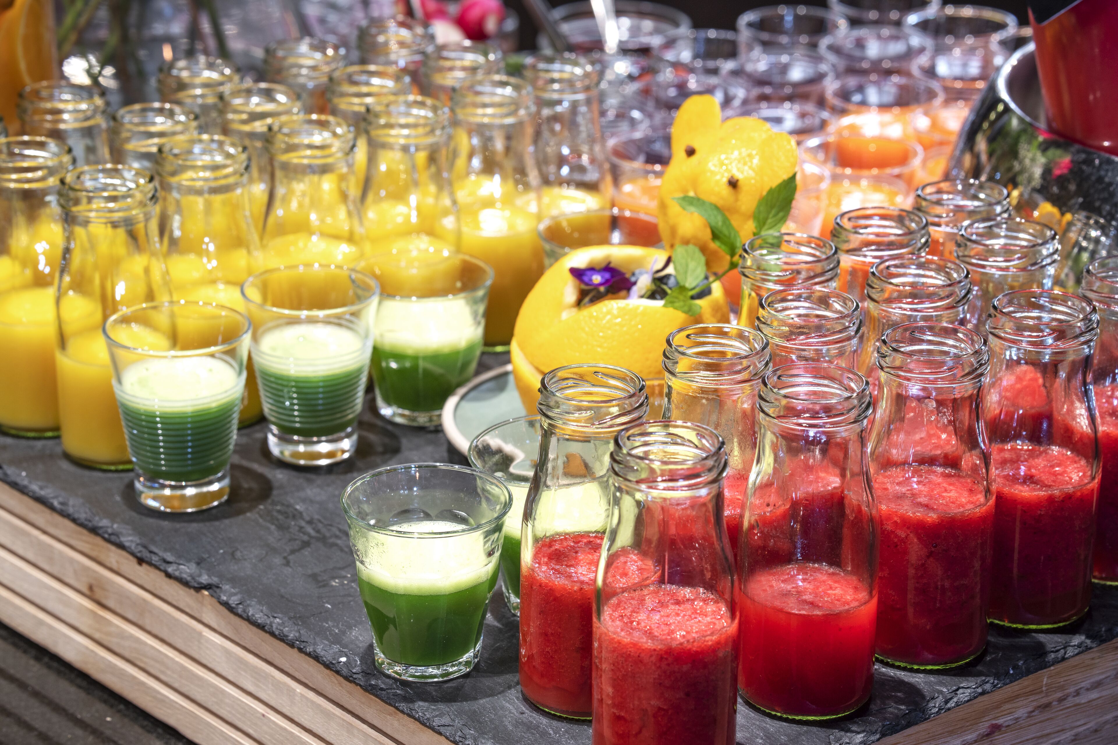 A variety of colorful smoothies in glass bottles and small glasses on a slate countertop.