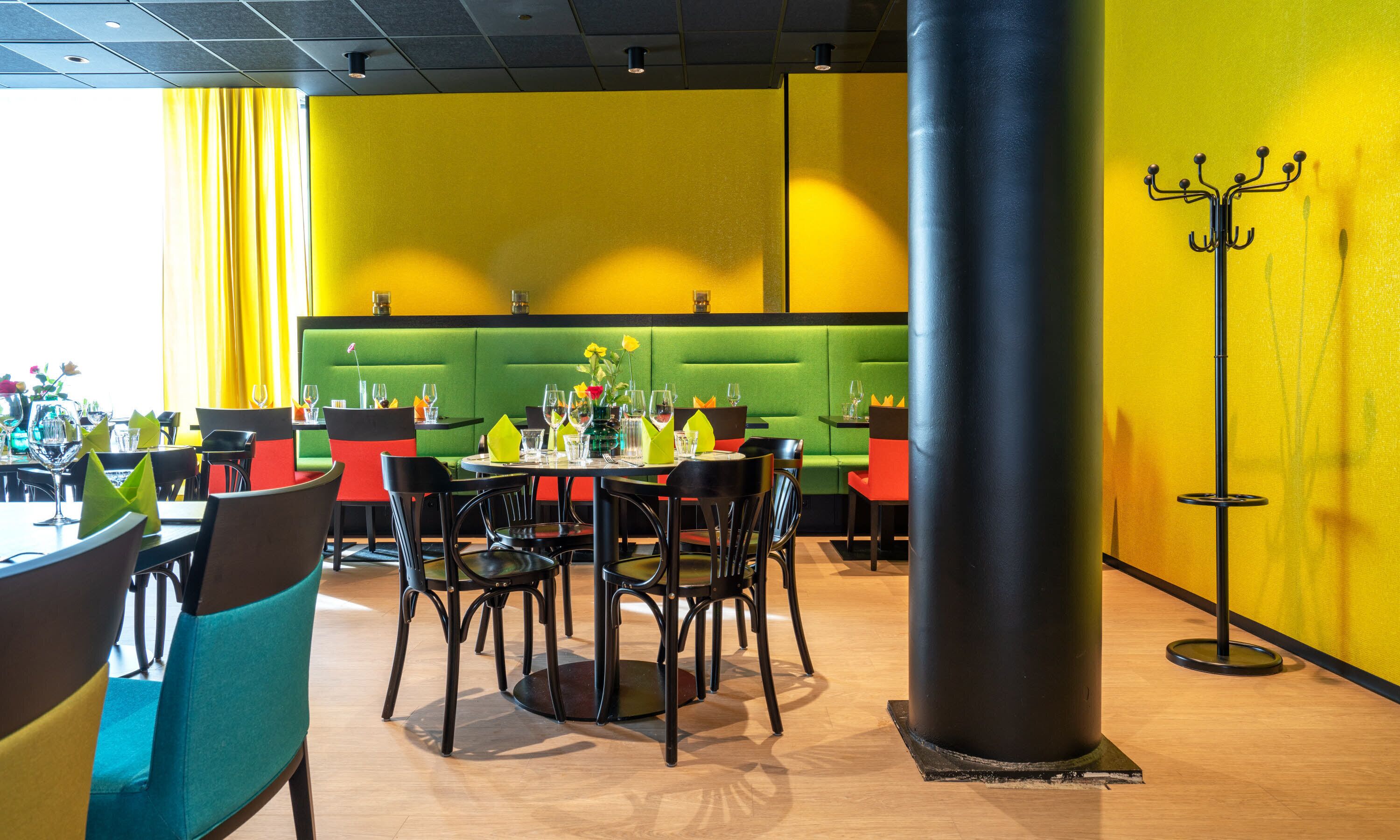 Un restaurante con mesas revestidas de verde, sillas negras y rojas, y paredes amarillas.
