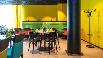 Un restaurante con mesas revestidas de verde, sillas negras y rojas, y paredes amarillas.