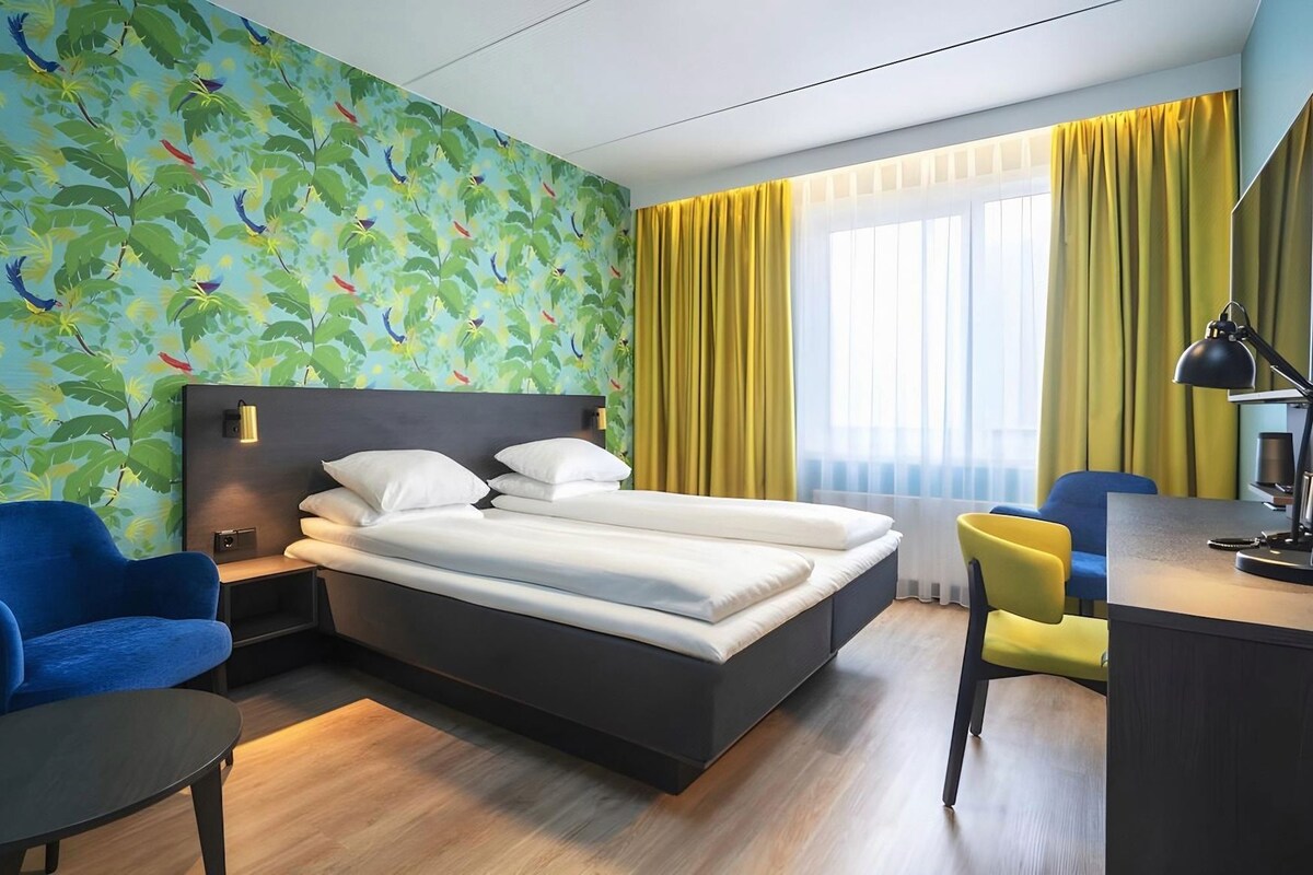 Une chambre d’hôtel avec un lit, un bureau, une chaise et un mur orné d’un motif végétal.