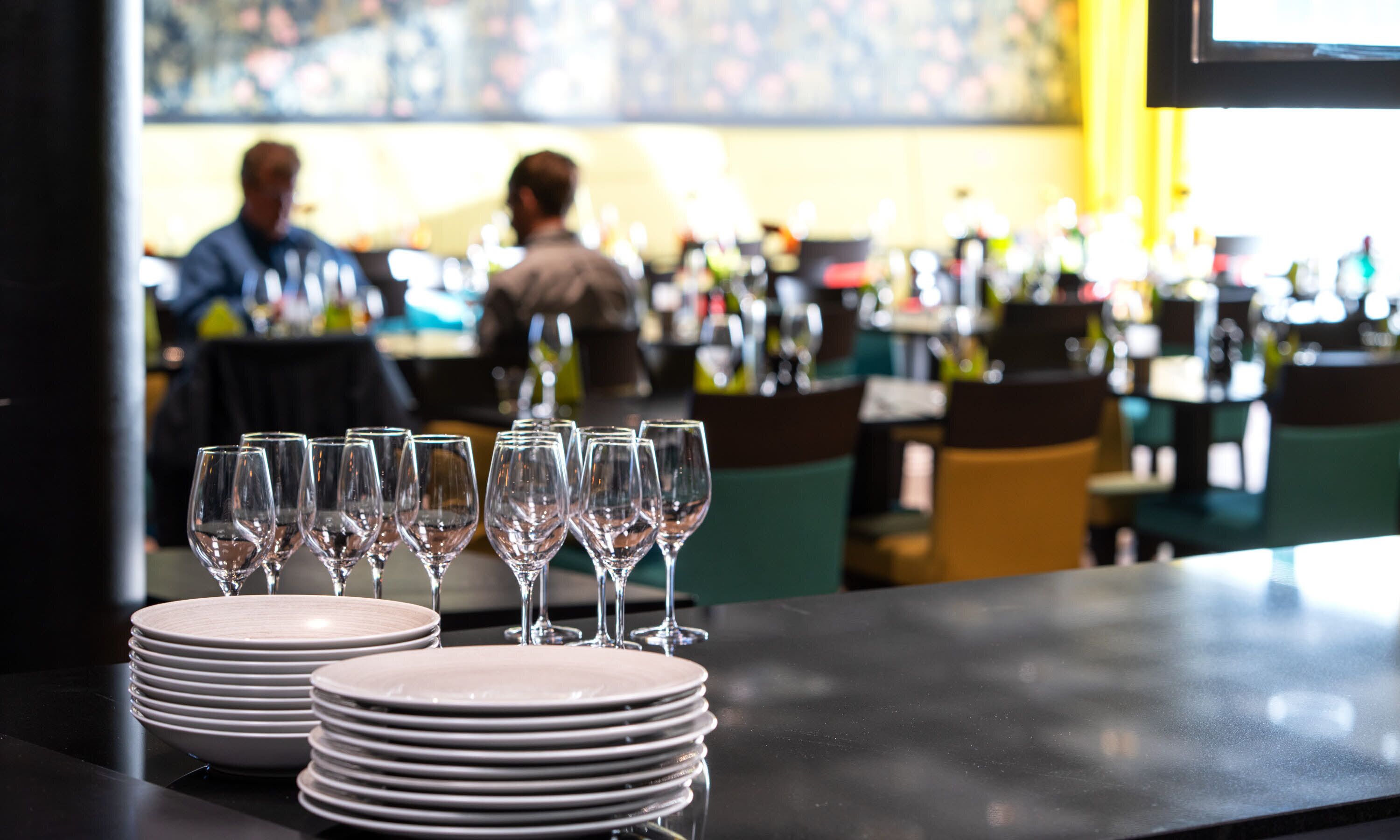 A bar area with stacked plates and wine glasses, a bar counter, and a seating area with chairs.