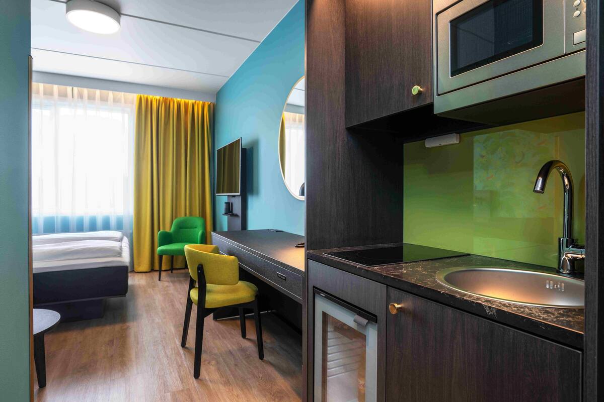 A compact hotel room with a bed, a desk, a chair, a microwave, a sink, and a television.