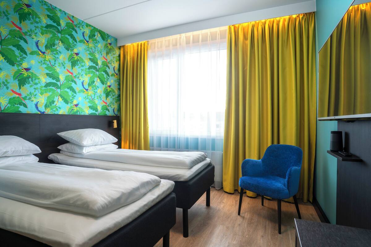 A hotel room with two beds, a blue chair, a TV, and a window with yellow curtains.