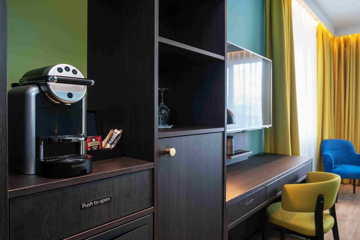 A compact room with a dark wood cabinet, a Nespresso coffee machine, a small shelf with a wine glass, and a yellow chair.