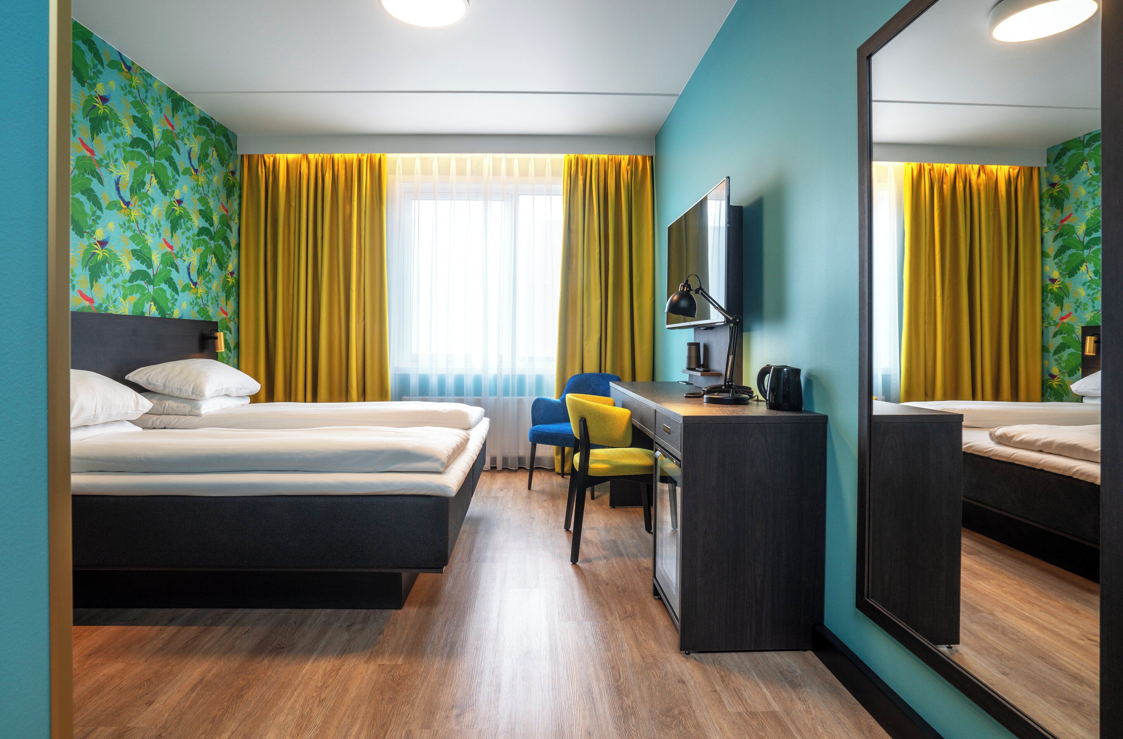 A hotel room with two beds, a desk, a chair, a mirror, and a window with yellow curtains.