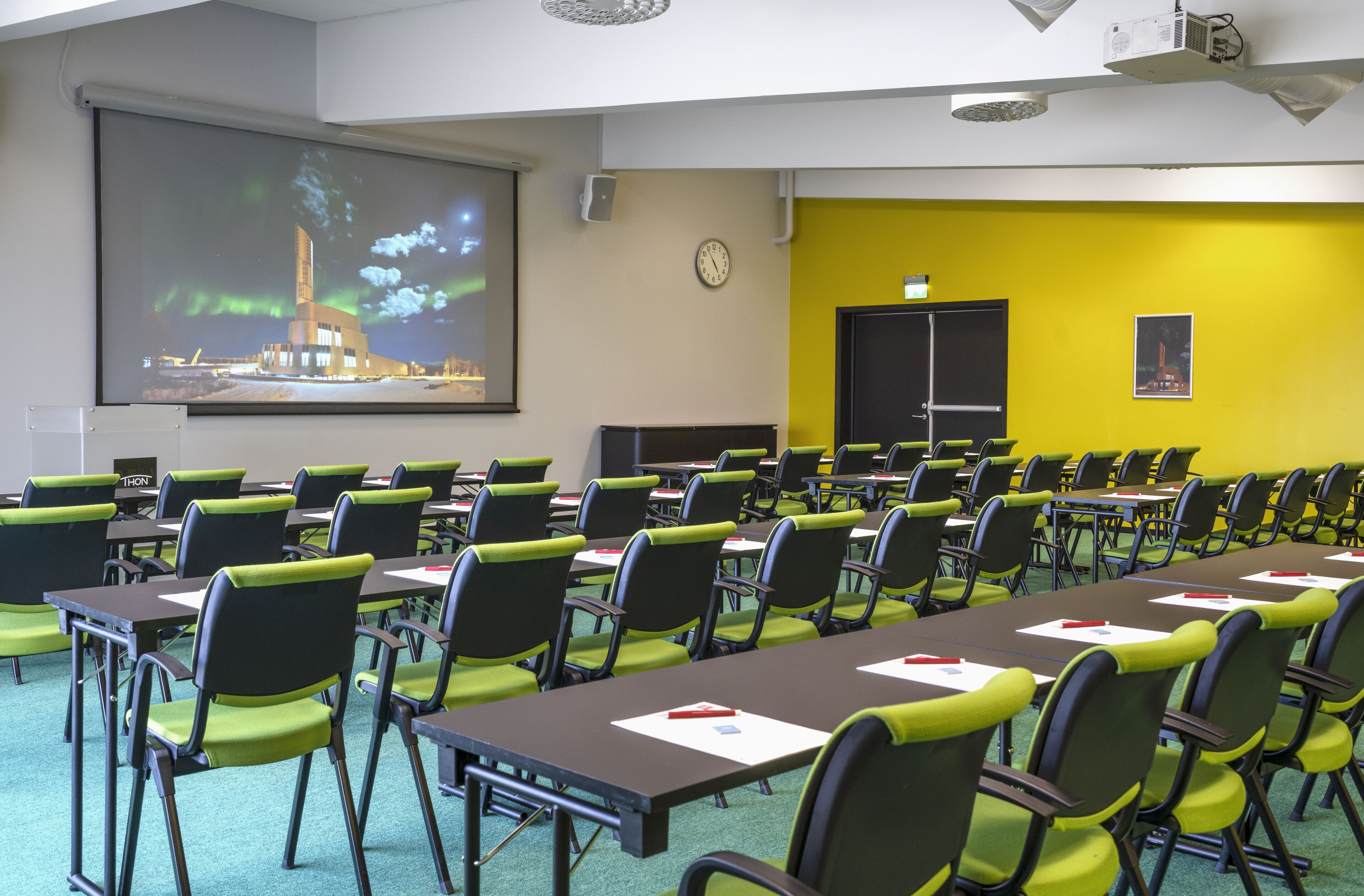 A conference room with rows of tables and chairs, a projector screen, and a clock on the wall.