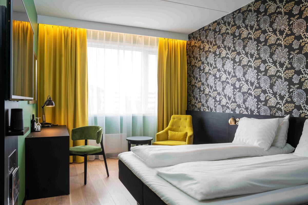 Quarto de hotel com cama, escrivaninha, cadeira, mesinha e uma grande janela com cortinas amarelas.