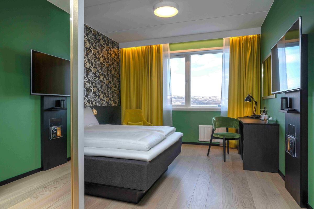 Quarto de hotel com cama, escrivaninha, cadeira, televisão e janela com cortinas.