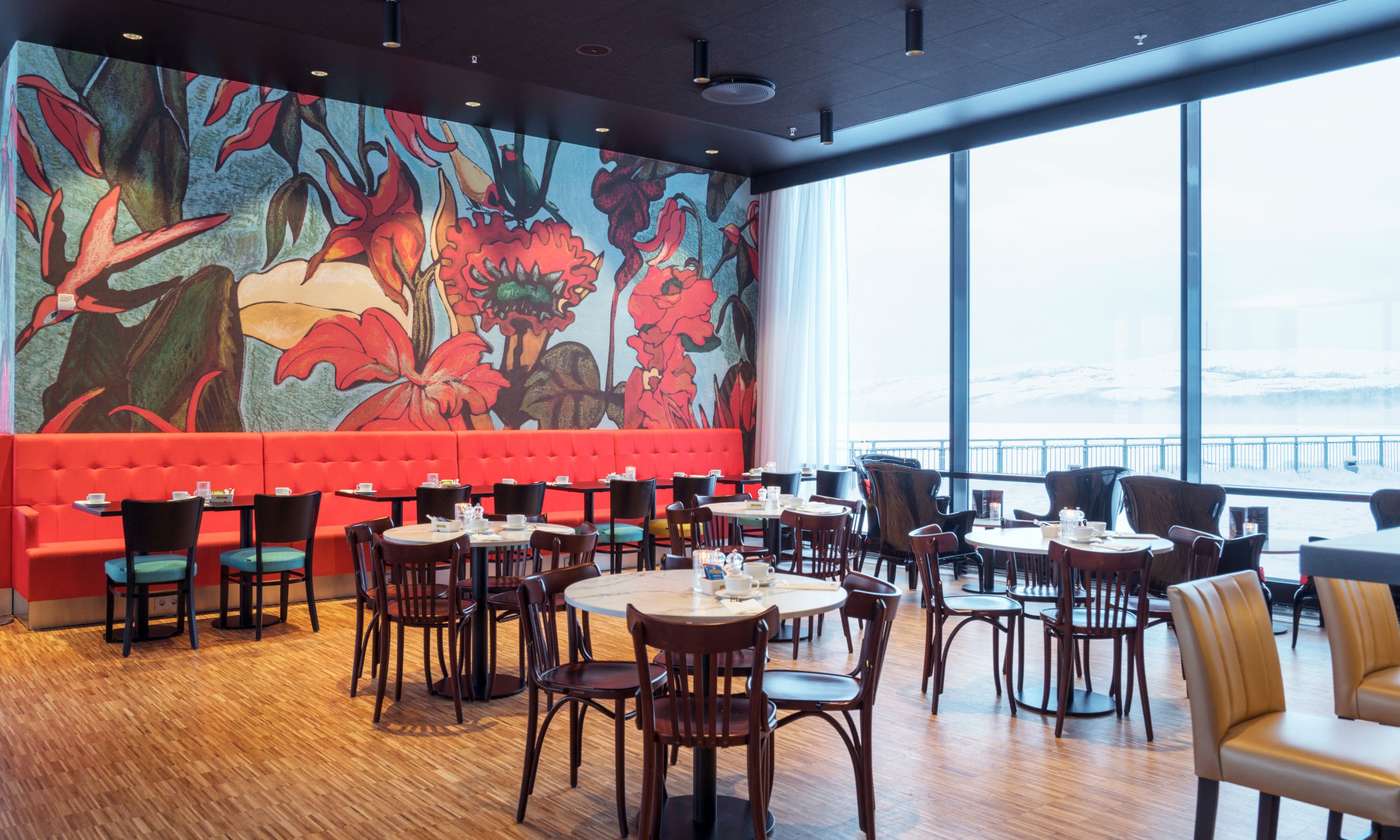 A restaurant with a vibrant floral mural, red seating, and a view of a snowy landscape through large windows.