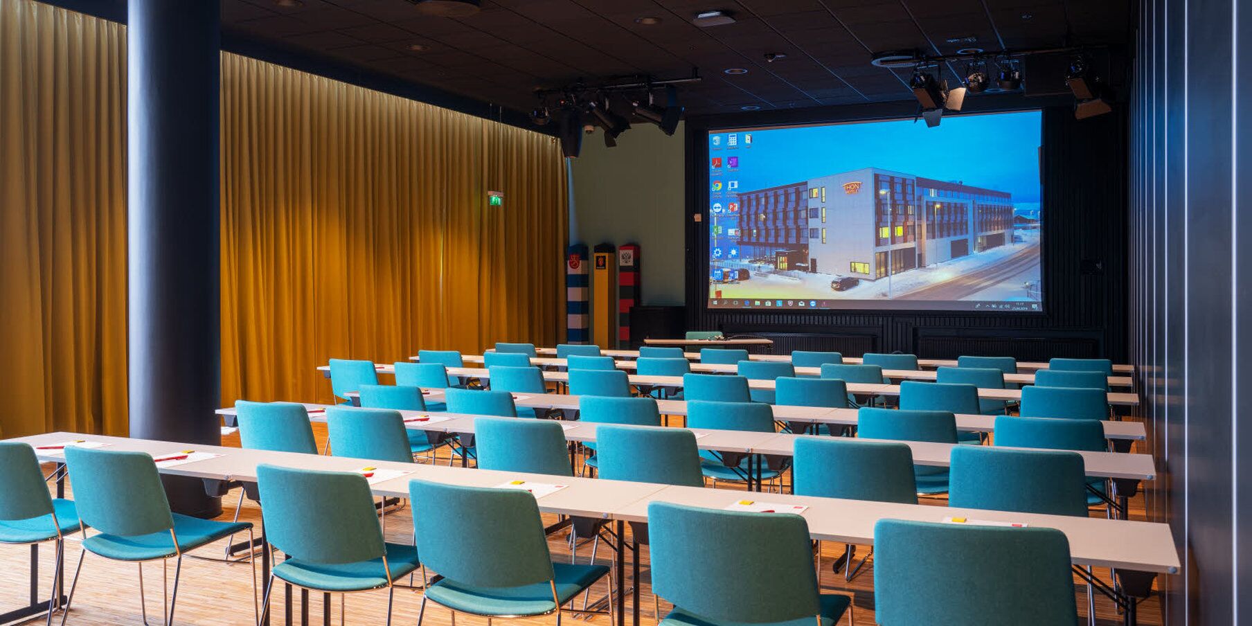 A conference room with rows of teal chairs and tables, a large screen displaying a building, and a curtain backdrop.
