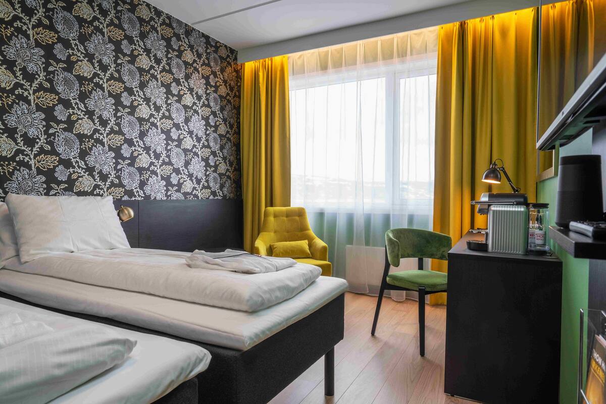 Quarto de hotel com cama, escrivaninha com luminária, cadeira e janela com cortinas.
