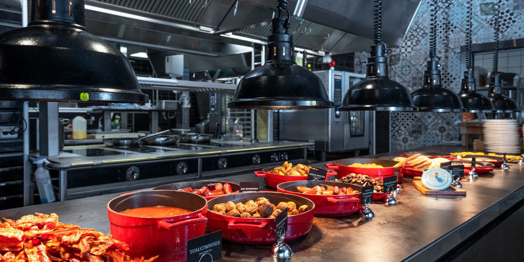 Ein Buffet mit verschiedenen Speisen in roten und schwarzen Behältern, ein Herd und industrielle Deckenleuchten.