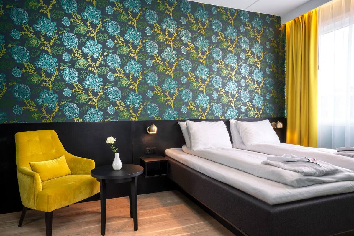 Habitación de hotel con una cama, un sillón amarillo, una mesita con un florero y un papel tapiz estampado.